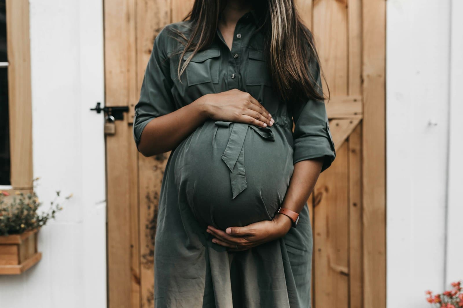 A pregnant woman in a sage green dress holding her bump, standing by a wooden door