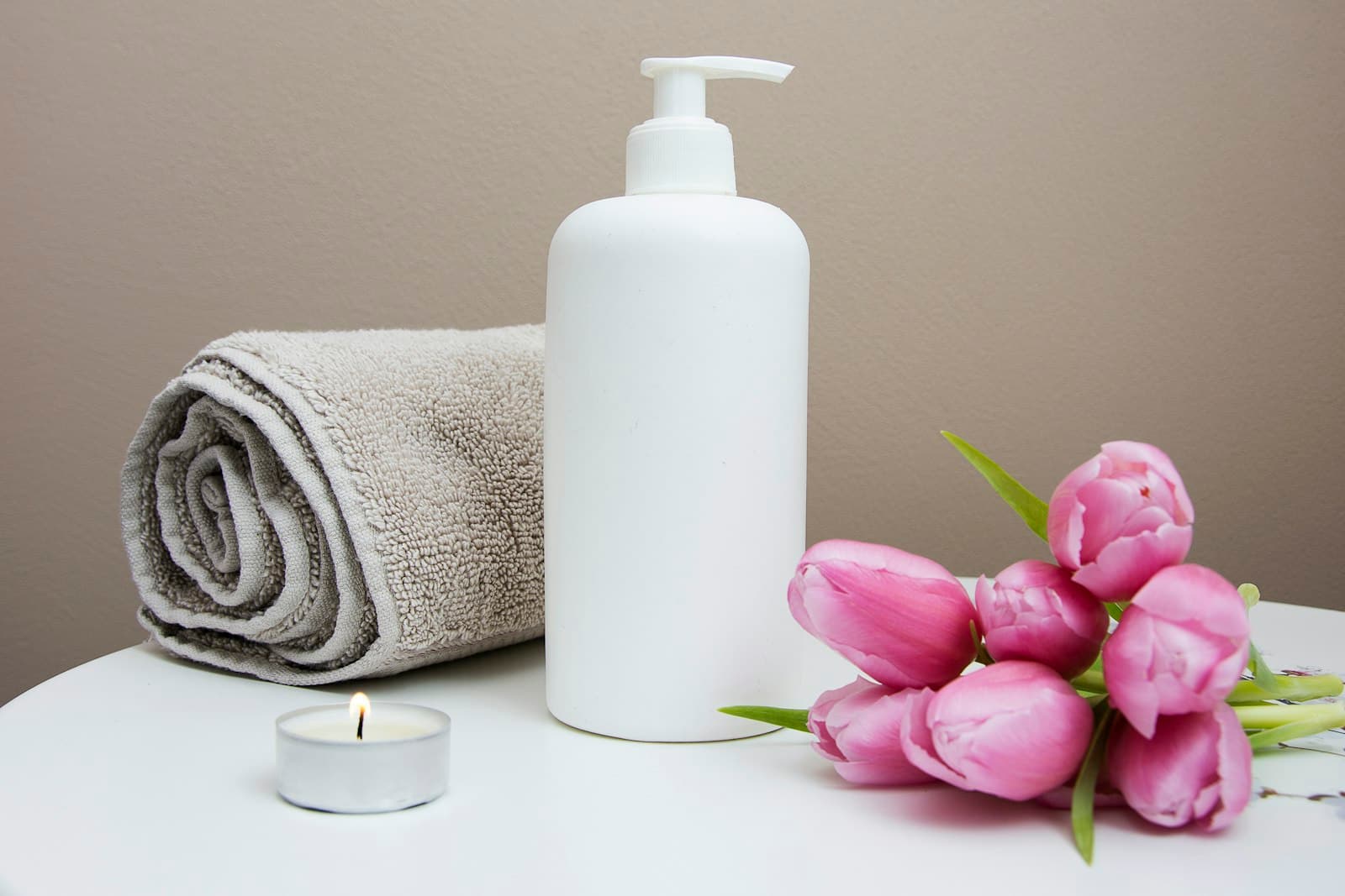 A rolled towel, massage lotion and a lit candle on a calm white table